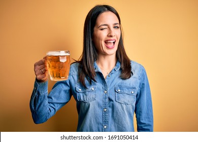 Young woman with blue eyes drinking jar of beer standing over isolated yellow background winking looking at the camera with sexy expression, cheerful and happy face.