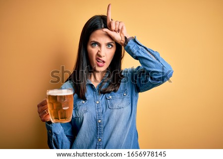 Young woman with blue eyes drinking jar of beer standing over isolated yellow background making fun of people with fingers on forehead doing loser gesture mocking and insulting.