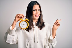 Young Woman With Blue Eyes Holding Alarm Clock Standing Over Isolated White Background Very Happy Pointing With Hand And Finger To The Side