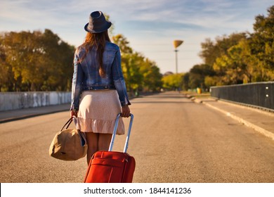 Young Woman With A Blue Hat, Denim Jacket, Skirt, Red Suitcase And Brown Bag Walking On A Road To Start A Journey Or A New Life, View From The Back.