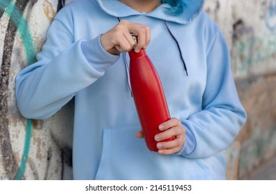 Young Woman In Blue Hoodie With Red Water Bottle Staying Outdoors Near Graffiti Wall. Clothing And Reusable Bottle Mockup.  Hipster And Adolescence Concept