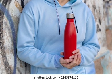 Young Woman In Blue Hoodie With Red Water Bottle Staying Outdoors Near Graffiti Wall. Clothing And Reusable Bottle Mockup.  Hipster And Adolescence Concept