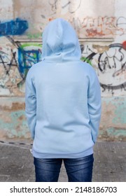 Young Woman In Blue Hoodie Stand With Her Back To Camera.  Adolescent Girl Stays Outdoors In Front Of Graffiti Wall. Clothing Mockup 