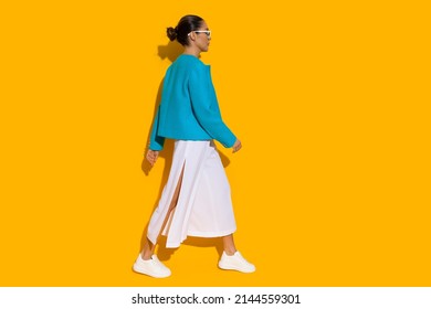 Young Woman In Blue Jacket, White Baggy Pants And Sneakers Is Walking. Side View. Full Length Studio Shot On Yellow Background.