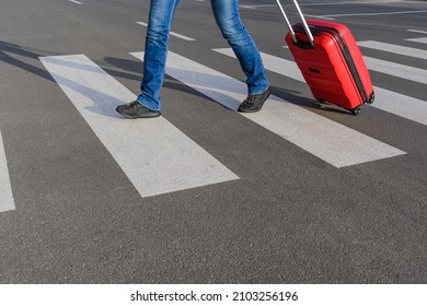 The young woman in blue jeans with red rolling bag is crossing road. The lady in blue jeans and black boots is walking by crosswalk. This is close up photo.