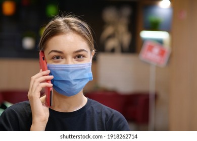 Young Woman In Blue Medical Protective Mask Talks On Smartphone Inside A Restaurant. Covid Concept Health And Safety, Coronavirus Quarantine, Social Distancing, Next Wave Covid Virus Protection