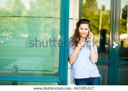 young woman in blue shirt standing in front of big glass door and using her mobile phone
