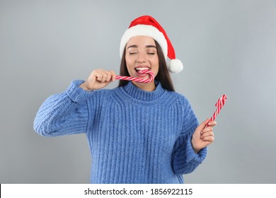Young woman in blue sweater and Santa hat eating candy canes on grey background. Celebrating Christmas