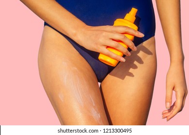 young woman in a blue swimsuit holding a plastic orange package spray bottle and putting sunscreen on her body. summer sun ultraviolet protection SPF on the beach pink background in studio