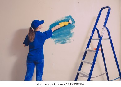 A Young Woman In A Blue T-shirt In Yellow Rubber Gloves And Cap Is Painting A Wall In An Apartment, There Is A Staircase Nearby.