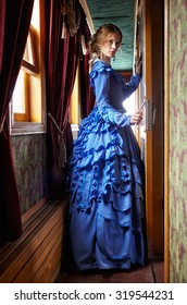 Young woman in blue vintage dress late 19th century standing near window in corridor of retro railway vehicle