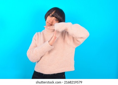 Young woman with bob haircut wearing pink knitted sweater shirt over blue wall feels tired and bored, making a timeout gesture, needs to stop because of work stress, time concept.