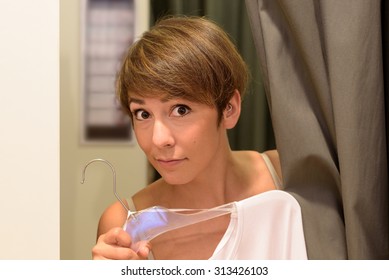 Young woman in a boutique fitting room peering out from behind the curtain in her bra as she holds up a top on a hanger in front of her