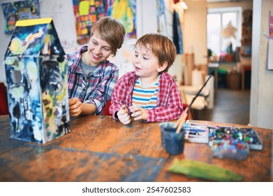 Young woman and boy sitting at a wooden table in an art studio, discussing a painted cardboard house. Colorful artwork displayed in the background - Powered by Shutterstock