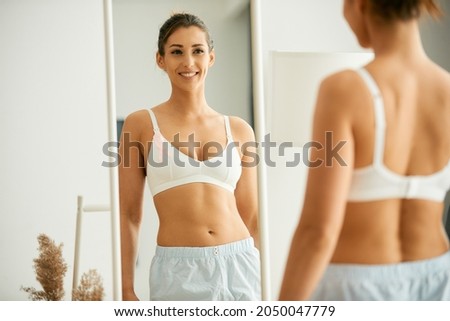 Young woman with breast cancer ribbon on a bra feeling satisfied while looking herself in a mirror at home.