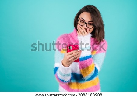 A young woman in a bright multicolored sweater on a blue background with glasses, holds the phone, laughs cheerfully, read a joke, anecdote, covers her mouth with her hand, laughs nonstop