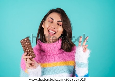A young woman in a bright multi-colored sweater on a blue background enjoys every piece of a delicious bar of airy milk chocolate, playfully sticks out her tongue