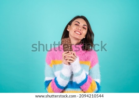 A young woman in a bright multi-colored sweater on a blue background enjoys every piece of a delicious bar of airy milk chocolate