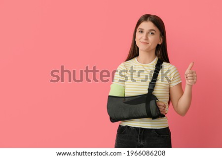 Young woman with broken arm showing thumb-up on color background