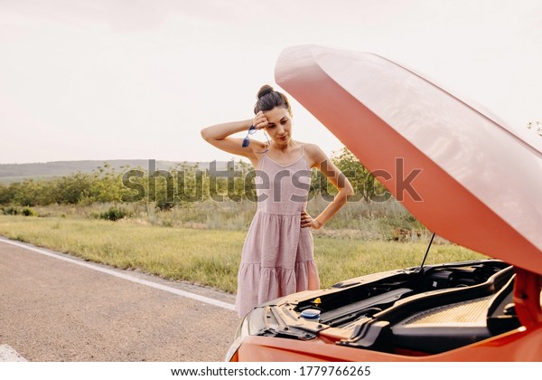 Young woman with a broken car on a highway, looking\
under the hood.