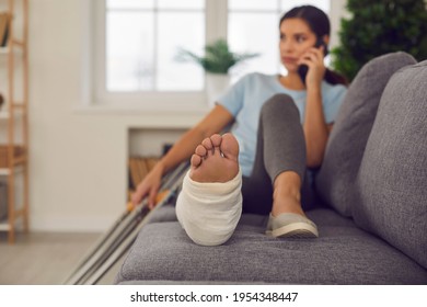 Young Woman With Broken Leg In Cast Lying Relaxing On Sofa With Crutches Nearby And Talking On Phone At Home, Selective Focus. Injury, Trauma, Recovery, Rehabilitation Concept