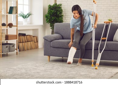 Young Woman With Broken Leg In Plaster Cast Trying To Stand Up From Sofa And Walk With Crutches In Living-room. Physical Injury, Bone Fracture, Car Or Home Accident, Rehabilitation Of People Concept