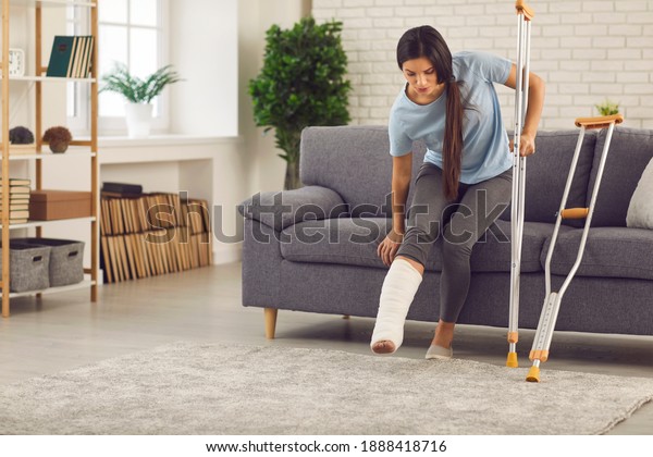 Young woman with broken leg in plaster cast\
trying to stand up from sofa and walk with crutches in living-room.\
Physical injury, bone fracture, car or home accident,\
rehabilitation of people\
concept