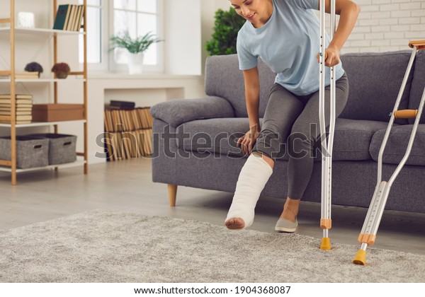 Young woman with broken leg in plaster cast tries\
to stand up from sofa and walk with crutches at home. Physical\
injury in domestic or car accident, bone fracture and\
rehabilitation of people\
concept