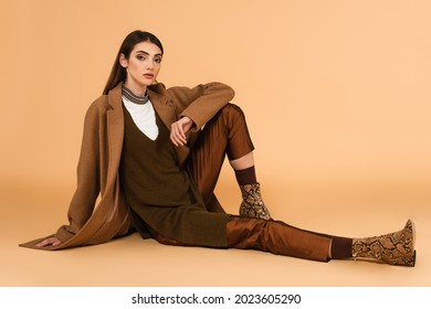 young woman in brown coat, satin pants and leather shoes sitting on beige background