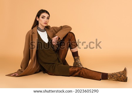 young woman in brown coat, satin pants and leather shoes sitting on beige background
