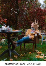 Young Woman Brown Coat Sits Chair At Table Reading Book With Plaid Thrown Over Her Head Open Air Against Autumn Reddened Foliage. Autumn Fashion Season. Rest, Tranquility, Backyard, Patio, Melancholy