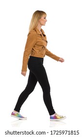 Young Woman In Brown Suede Leather Jacket, Black Jeans And Colorful Sneakers Is Walking. Side View. Full Length Studio Shot Isolated On White.