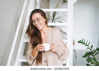 Young woman with brunette long hair in cozy knitted cardigan with cup of tea in hands in bright interior at home