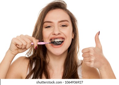 Young Woman Brushing Her Teeth With A Black Tooth Paste With Active Charcoal, And Black Tooth Brush On White Background, And Showing Thumbs Up