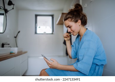 Young woman brushing her teeth and scrolling her smartphone, morning routine concept. - Powered by Shutterstock