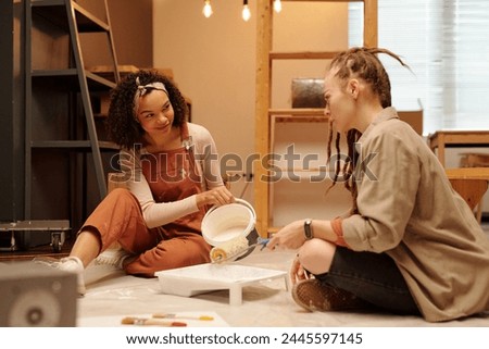 Young woman with bucket pouring white paint in tray while sitting on the floor and looking at girl paintroller during cafe renovation work