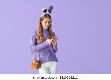 Young woman in bunny ears with basket of Easter eggs using mobile phone on lilac background - Powered by Shutterstock