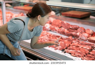 Young woman buyer of meat department of grocery store, looking at meat in refrigerator window. Female customer points with finger and orders beef brisket and sirloin - Powered by Shutterstock