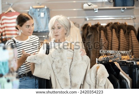 Young woman buyer in store has removed hanger with fur coat from window and lay heads together with old woman friend about purchasing. Girl imagine warmth that this coat will give on winter