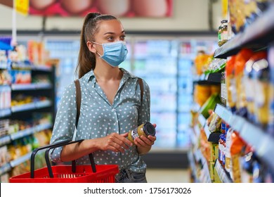 Young Woman Buyer Wearing Medical Protective Mask Among The Shopping Shelves Chooses Food Products In A Grocery Store 