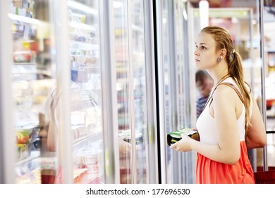 Young Woman Buying Dairy Or Refrigerated Groceries At The Supermarket In The Refrigerated Section Opening Glass Door Of The Fridge