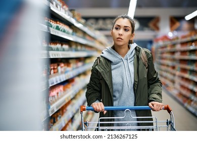 Young Woman Buying Groceries And Pushing Shopping Cart While Walking Among Produce Aisle In Supermarket.