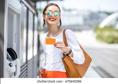 Young woman buying a ticket for public transport at the automatic machine or withdrawing money with a card standing outdoors at the modern city