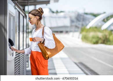 Young woman buying a ticket for public transport at the automatic machine or withdrawing money with a card standing outdoors at the modern city