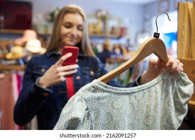 Young Woman Buying Used Sustainable Clothes From Second Hand Charity Shop Taking Picture Of Dress On Mobile Phone