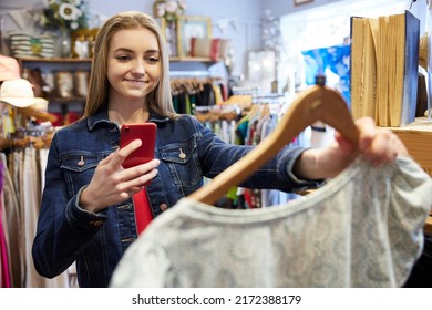 Young Woman Buying Used Sustainable Clothes From Second Hand Charity Shop Taking Picture Of Dress On Mobile Phone