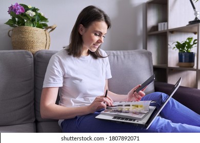Young woman buys goods, cosmetics, clothes online. In the hands of paper catalog laptop smartphone, female makes purchases at home, sitting on the couch