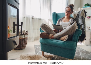 Young woman by the fireplace, sitting in a cozy armchair, with a warm blanket, using a tablet - Powered by Shutterstock