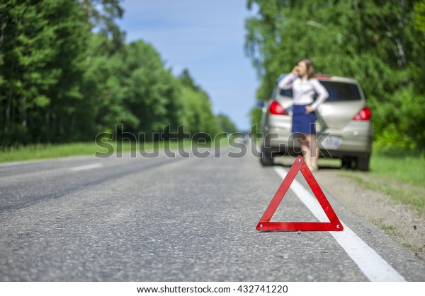 Young woman calling for car\
assistance after breakdown on the road. Focus on red triangle\
warning sign.
