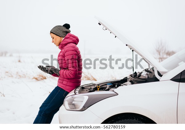 Young woman calling for help or assistance after\
her car breakdown in the winter. Broken down car with open hood on\
a country road.